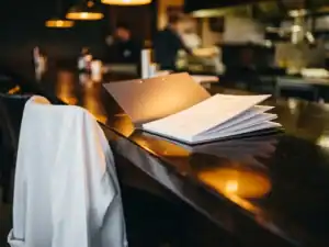Loonmap en arbeidscontract op restaurantbar, met wit koksjas over kruk en warme hanglampen op de achtergrond.