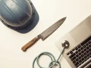 Bovenaanzicht van een koksmes, bouwhelm, stethoscoop en laptoptoetsenbord op wit oppervlak, symbolen van diverse sectoren.