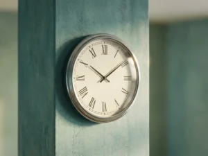 Analoog wandklok met chromen rand op een zachte teal betonnen muur in warm, diffuus daglicht.