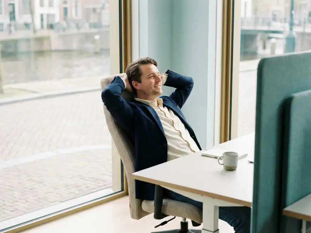 Ontspannen ondernemer in ergonomische stoel met handen achter het hoofd in een rustig, minimalistisch Nederlands kantoor met natuurlijk licht.