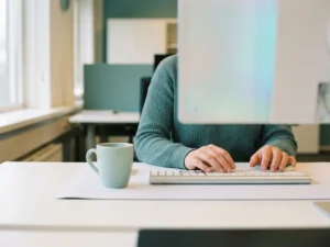 Handen van een salarisadministrateur boven een modern toetsenbord in een strak Nederlands kantoor, met een koffiekop en zacht monitorlicht.