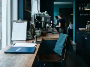 Horecabar met werkrooster, loonstrookmap en koffietamper, schort over barkruk in modern Nederlands restaurant met teal en marineblauw interieur.