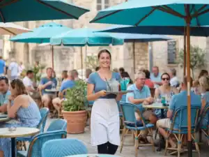 Seizoensmedewerker met wit schort draagt dienblad met drankjes op zonnig terras met parasols en caféstoelen.